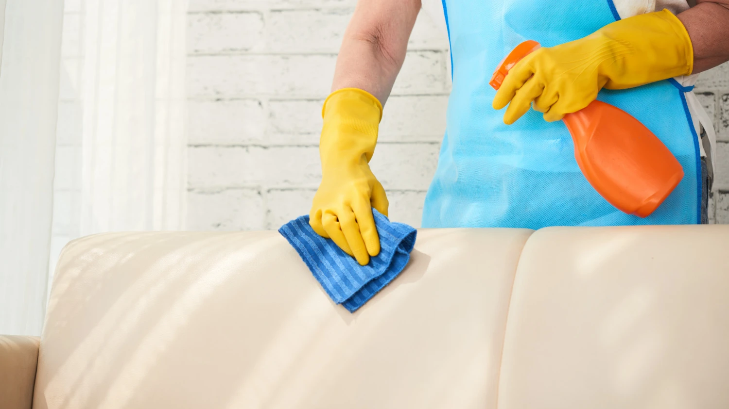 Cleaner in yellow gloves wiping a beige leather sofa with a cloth and spray bottle.