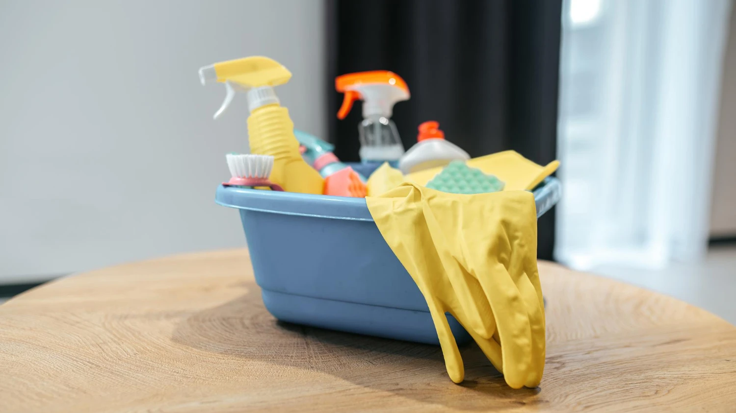 Blue basket with cleaning supplies and yellow rubber gloves on a wooden table.